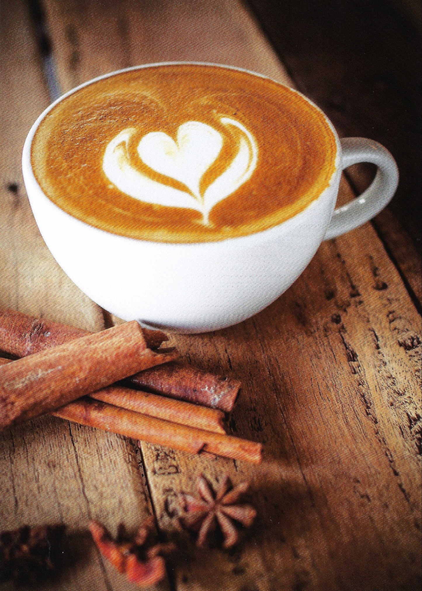 A postcard featuring a steamed milk heart design in a cup of cappuccino, surrounded by spices like cinnamon sticks and aniseed on a wooden surface.