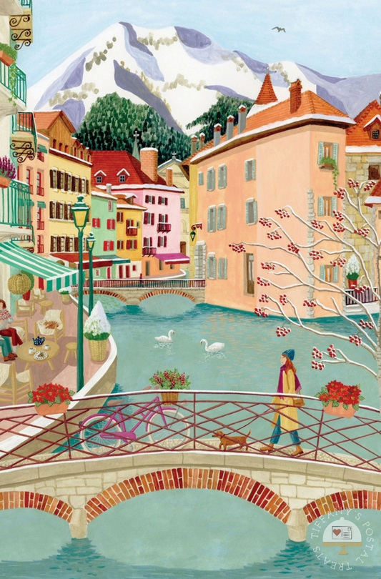 Annecy Postcard by Mélanie Voituriez featuring an illustration of a European village scene with pastel buildings, a canal, stone bridges, swans, a person walking with a dog, and snow-capped mountains in the background.