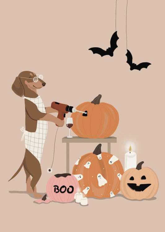 Halloween Postcard by Alfie's Greetings for Nobis Design featuring a brown dog wearing glasses and a white apron, carving pumpkins. The scene includes a pink pumpkin with 'BOO' written on it, a carved jack-o'-lantern, a lit candle, hanging bats, a glass of red wine and a warm beige background.