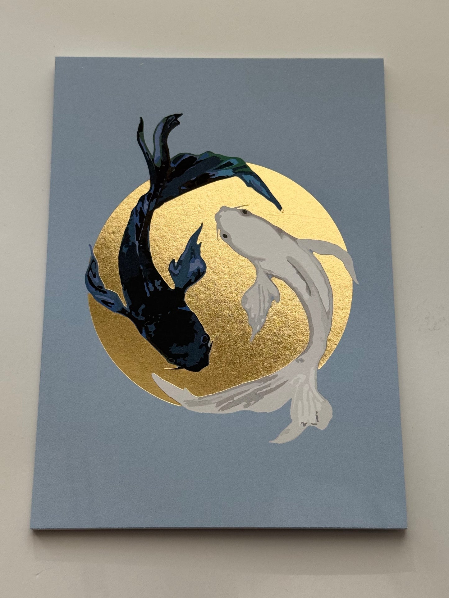 Koi Fish Yin Yang Postcard by Nobis Design featuring koi fish with one black fish and one white fish swimming in front of a large gold circle on a muted blue-gray background