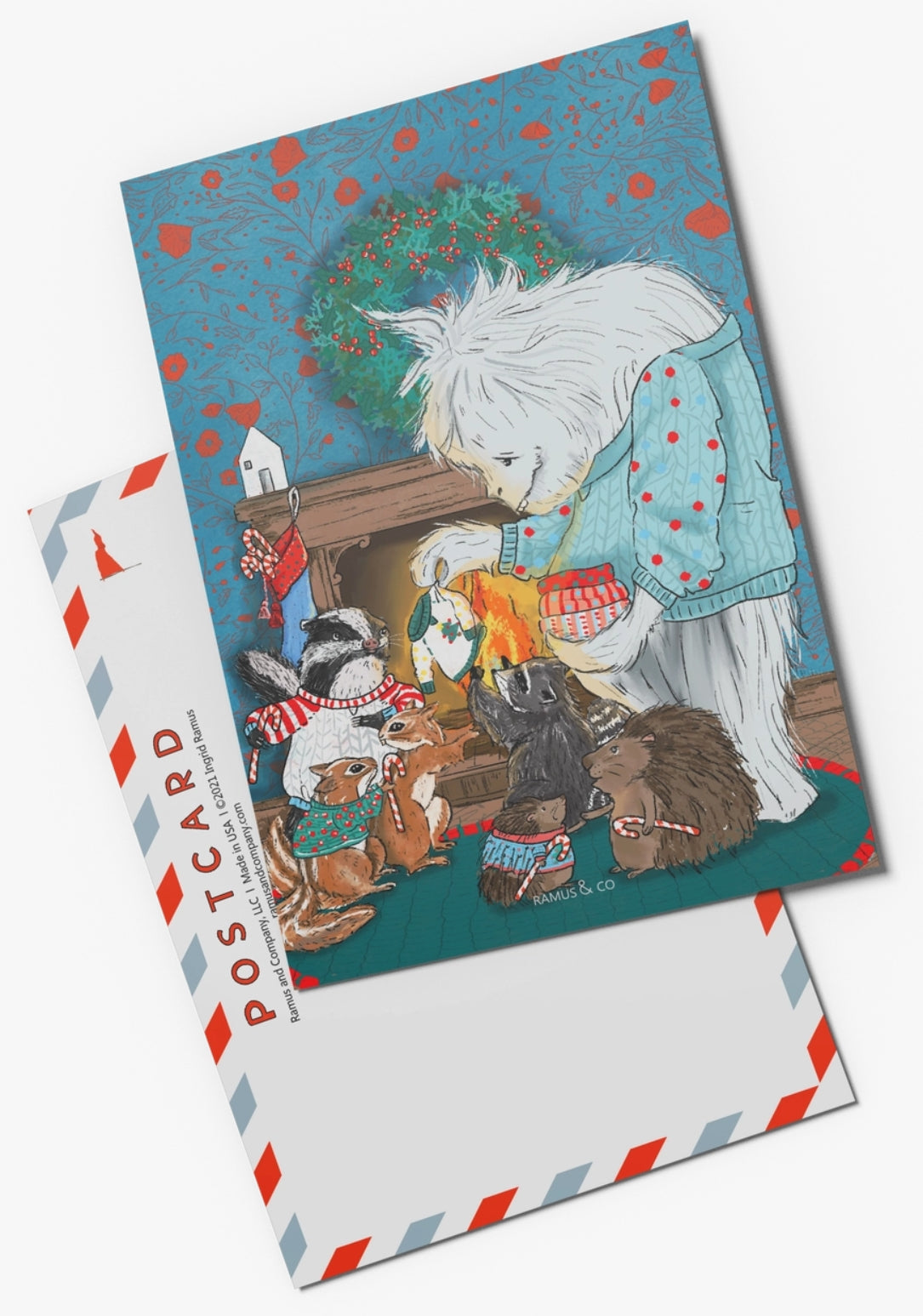 Yeti Sweater Club Postcard by Ramus and Company featuring a white Yeti  handing out Christmas Sweaters to several small animals around a fireplace. The background includes a blue patterned wall, a green wreath with red berries, stockings, and holiday decorations with the postcard back behind it.